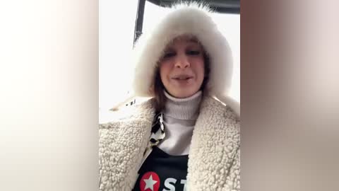 Media: Video of a person wearing a white fur hat, a white turtleneck sweater, and a beige fur coat, with a red and white graphic tee peeking out, inside a vehicle.