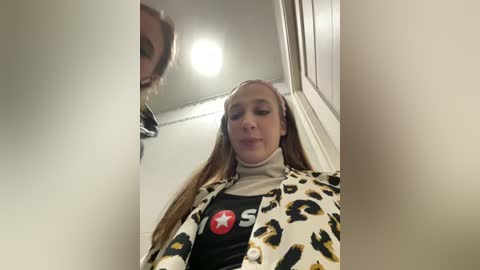 Media: Video of a young woman with long brown hair, wearing a leopard-print jacket over a black t-shirt with a red star and white text, inside a narrow, brightly lit hallway.