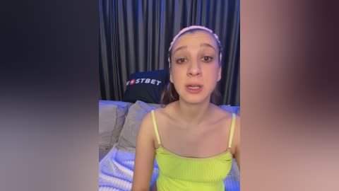 Media: A video of a young Caucasian woman with light skin and brown hair, wearing a yellow tank top and headband, sitting on a bed with gray sheets.