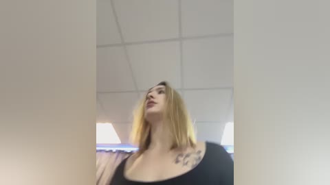 Media: A video of a blonde woman with a large tattoo on her shoulder, wearing a black top, looking up in an office with fluorescent lights and white ceiling tiles.