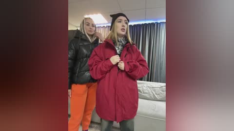 Media: Video of two women in casual attire, one in a red coat, standing in a living room with a beige couch and gray curtains.