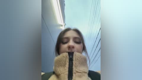 Media: A video of a young woman with long brown hair, wearing a beige fur-lined jacket, looking down, taken from a low angle in a narrow corridor with blue walls and overhead fluorescent lights.