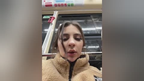 Media: Video of a young woman with brown hair and fair skin, wearing a beige fleece jacket, seated in a subway car with a reflective surface behind her.