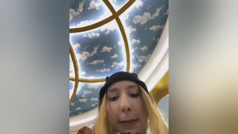 Media: A video of a young Caucasian woman with long blonde hair, wearing a black hat and a yellow top, standing beneath a whimsical sky mural with clouds and a sun.