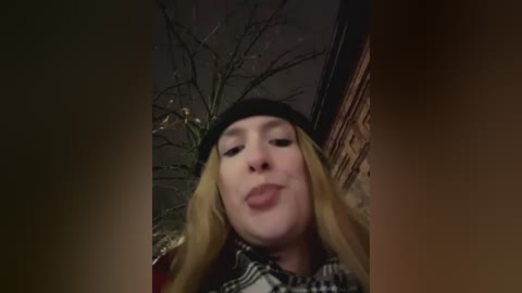 Media: Video of a blonde woman with fair skin, wearing a black beanie and checkered shirt, making a pouty face, against a dark, leafless tree and brick wall background.