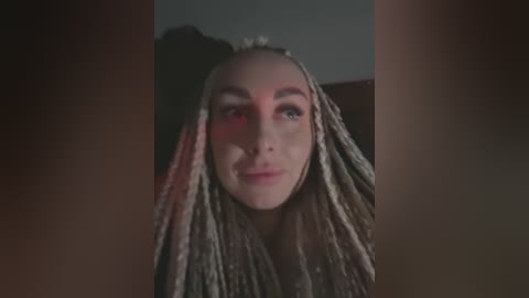 Media: A close-up video of a young woman with light skin and long, braided dreadlocks, wearing minimal makeup. The background is dark, with a faint red light casting a warm glow on her face.