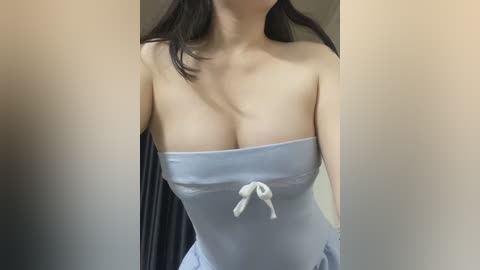 Media: Video of an Asian woman with fair skin, wearing a strapless light blue dress with a white bow, standing in a room with beige walls and dark curtains.