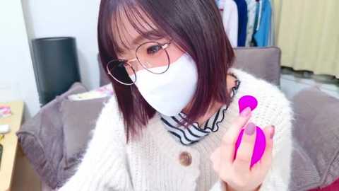 Video of a young woman with shoulder-length brown hair, glasses, and a white face mask, wearing a cream knit cardigan and a black and white striped shirt. She has bright pink nails and is holding a pink phone. Background features a gray couch and a cluttered room.