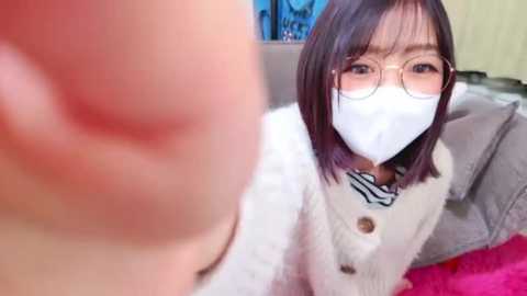 Media: Video of an East Asian woman with shoulder-length dark hair, wearing glasses and a white face mask, sitting on a couch, taking a selfie with a blurred man in the foreground.