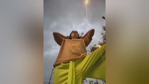 Media: A video of a young person, possibly a boy, with a light brown complexion, wearing a light orange hoodie and bright green pants, arms raised, against a cloudy sky background.