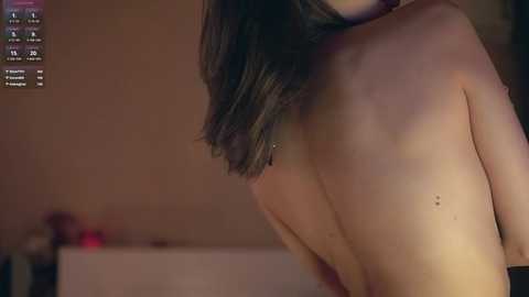 Media: Video of a slender, light-skinned woman with straight, shoulder-length dark hair, seen from the back, topless, standing in a dimly lit, beige-walled room with a blurred background.