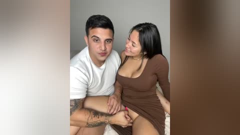 Media: A video shows a tattooed man in a white T-shirt and a woman in a brown dress, seated on a bed. They're smiling and holding hands.