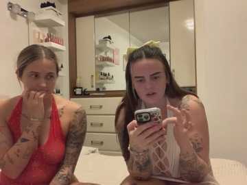 Media: Video of two women with tattoos, one in a red halter top, the other in a white lace top, sitting in a modern bathroom with white cabinets and a mirror.