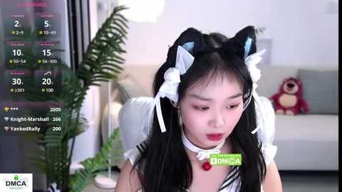 Media: Video of a young East Asian woman in a white maid outfit with black cat ears and tail, sitting in a modern living room.