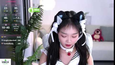 Media: Video of an East Asian woman in a cat maid cosplay, with black hair in twin tails, white and black outfit, large red choker, and cat ears. Background features a modern living room with a green plant, plush toys, and a white sofa.