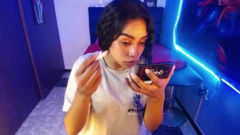 Media: Video of a young woman with short black hair, wearing a white T-shirt, looking at a smartphone while sitting on a tiled floor in a dimly-lit room with blue neon lighting.