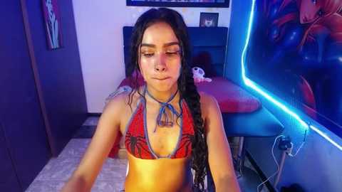 Media: Video of a young Latina woman with long black hair in a red bikini, sitting in a dimly lit room with neon lights and colorful decor.