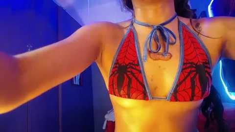 Media: Video of a woman with medium skin tone wearing a red and black spider web bikini with a halter neck and a blue rope choker, taken indoors with blue lighting and a mirror in the background.