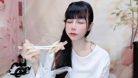 Media: Video of a fair-skinned woman with long black hair and bangs, wearing a white top, eating noodles with chopsticks in a modern, minimalistic room with a potted plant and abstract art.