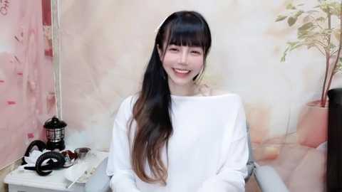 Media: A video of a young woman with long, straight, dark hair, wearing a white off-the-shoulder sweater, smiling, sitting in front of a pastel-colored wall with a plant and a black lantern.