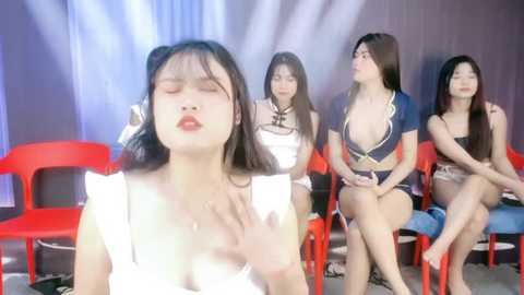 Media: Video of four Asian women with different hair and makeup styles, sitting on red plastic chairs, looking somber, in a dimly lit room.