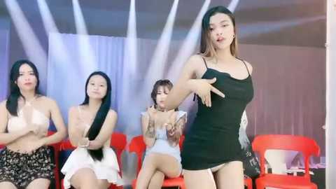 Media: Video of four Asian women, one in a black dress, dancing energetically in a dimly lit room with red chairs and white curtains.