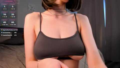 Media: A video of a woman with a medium build and short brown hair, wearing a tight, dark gray sports bra that accentuates her large breasts, taken in a dimly lit bedroom with grey curtains.