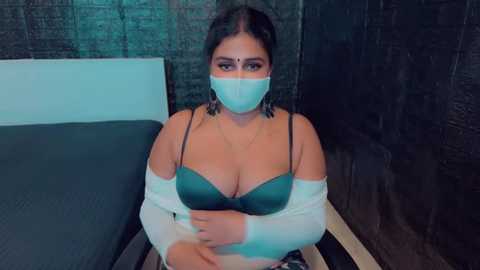 Media: Video of a woman with light brown skin, wearing a teal bra, white face mask, and a white cardigan, seated on a bed with dark gray curtains in the background.