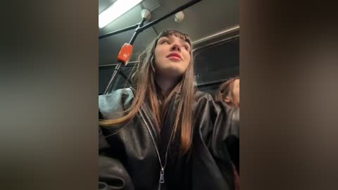 Media: A video of a woman with long brown hair in a black leather jacket, seated in a dimly lit train car. She has a neutral expression and is looking upwards.