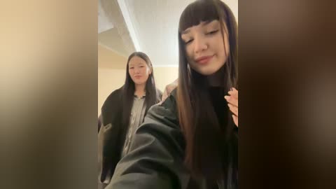 Media: Video of two young Asian women, one with long straight black hair, wearing a black leather jacket, and the other with bangs and a black shirt, standing in a dimly lit corridor.