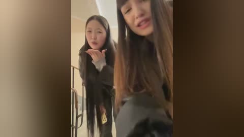 Media: Video of two young Asian women, one with long hair and a black coat, blowing a kiss, standing in a dimly lit hallway with beige walls.