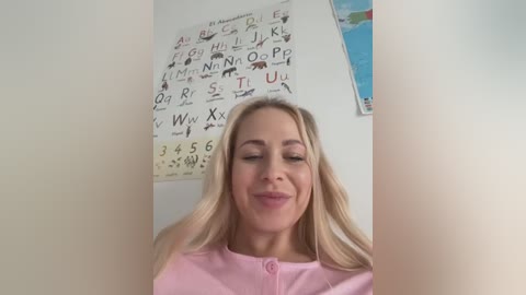 Media: Video of a smiling blonde woman with long hair, wearing a pink blouse, standing in a classroom with educational posters and a world map on the wall.