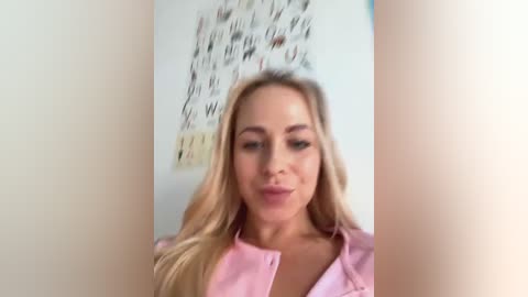 Media: Video of a Caucasian woman with long blonde hair, fair skin, wearing a pink shirt, standing in a blurred room with abstract art on the wall.