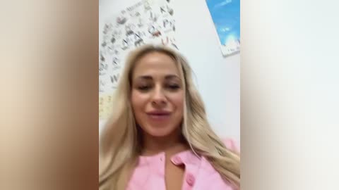 Media: A video of a smiling, fair-skinned blonde woman with long hair, wearing a pink blouse, in a white room with a blue wall calendar and a poster.