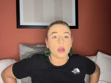 Media: Video of a fair-skinned woman with blonde hair, wearing a black t-shirt, green hoop earrings, and sitting on a white couch against a red wall with a framed beige abstract painting.