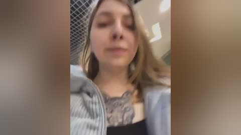 Media: A blurry, out-of-focus video of a young woman with fair skin, wearing a gray sweater and a hat, in a bathroom with a tiled wall and a mirror.