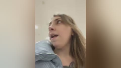 Media: A blurred video shows a young woman with long brown hair, wearing a blue denim jacket, leaning against a beige wall, mouth slightly open, appearing surprised or in thought.