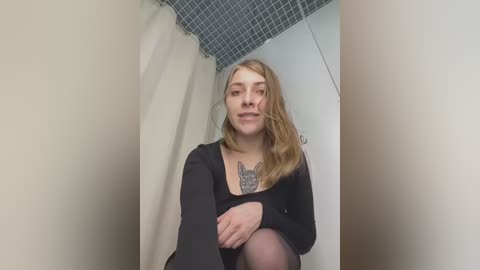 Media: A video of a young woman with fair skin, long blonde hair, and a black top featuring a large tattoo of a wolf on her chest. She sits in a small, beige-tiled bathroom with a blue-tiled ceiling.