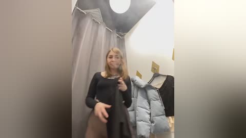 Media: Video of a young woman with light skin and straight, blonde hair, wearing a black dress and a blue puffer jacket, standing in a fitting room with beige walls and a black curtain.