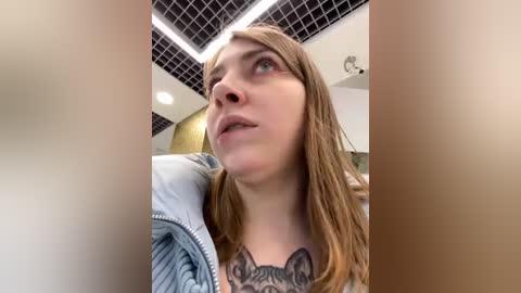 Media: Video of a young woman with long, light brown hair, fair skin, and a large tattoo of a cat on her chest, looking upward in an indoor setting with a grid ceiling.