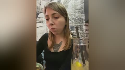 Media: Video of a young woman with long, straight blonde hair, wearing a black top, sitting at a table with a glass jar of lemonade and a straw. She has a tattoo of a fox on her chest.