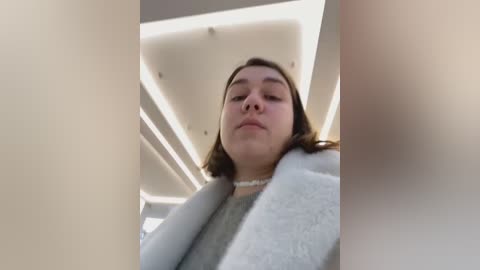 Media: A video of a young woman with medium-length brown hair, wearing a white pearl necklace and a gray coat, looking down. The background features a modern, light-colored ceiling with recessed lighting.