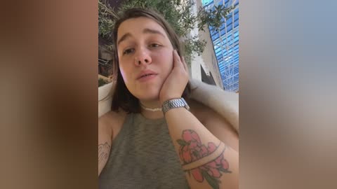 Media: Video of a young woman with shoulder-length brown hair, wearing a grey tank top, resting her chin on her hand, showing a colorful tattoo on her forearm. Background features a potted plant and a blue window frame.