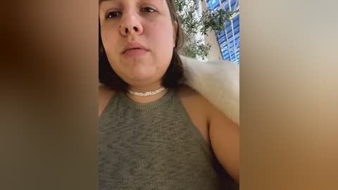 Media: A video of a young woman with medium skin tone, short dark hair, and a small nose ring, wearing a gray tank top and a pearl necklace, indoors with a blue tiled wall and green plants in the background.