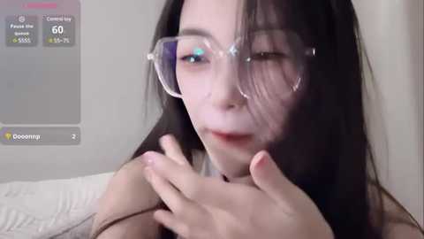 Media: Video of a fair-skinned Asian woman with long black hair, wearing large, clear glasses and a black top, touching her face. Background shows a white bed and a virtual overlay with statistics.