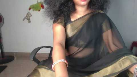 Media: Video of a woman with medium brown skin and curly hair, wearing a sheer black sari with gold trim. She sits on a black chair, hands visible. Background includes a plain wall, red shoes, and a green stuffed animal.