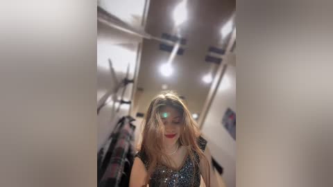 Video of a woman with long blonde hair, wearing a glittery black dress, standing in a dimly lit room with ceiling lights and hanging clothes in the background.