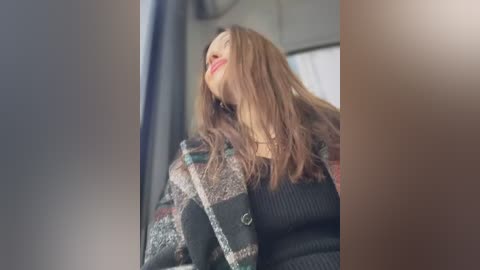 Video of a young woman with long, wavy brown hair, wearing a black sweater and patterned scarf, smiling and leaning against a car window.