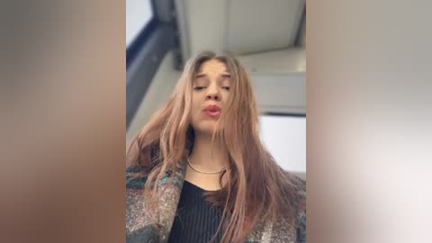 Video of a young woman with long, straight, light brown hair, wearing a black sweater and a patterned coat, blowing a kiss, indoors with a blurred, neutral background.