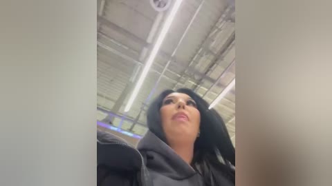 Media: A video captures a woman with a fair complexion, straight black hair, and wearing a dark hoodie, standing in a dimly lit, industrial setting with exposed pipes and fluorescent lights.